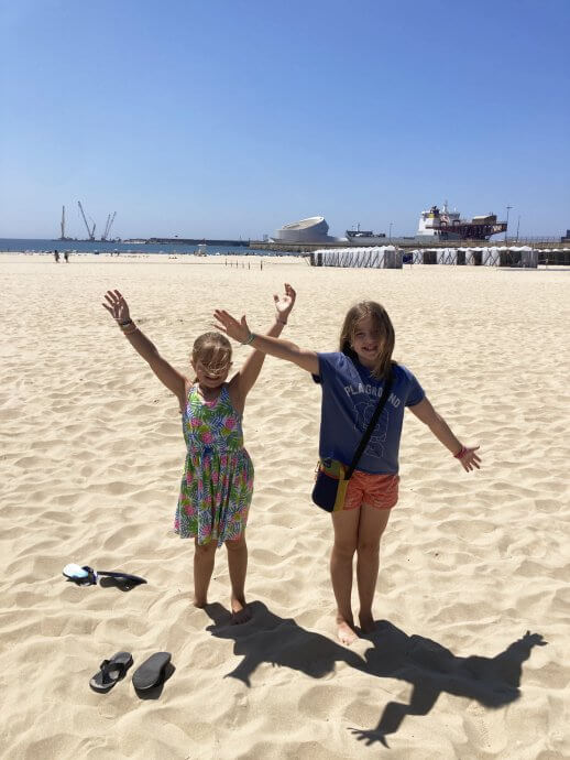 Two girls standing in the sand in Portugal with their arms up and flip flops in the sand, having just arrived.